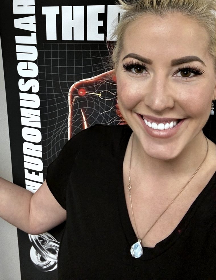 Elle smiling beside her Neuromuscular Therapy banner